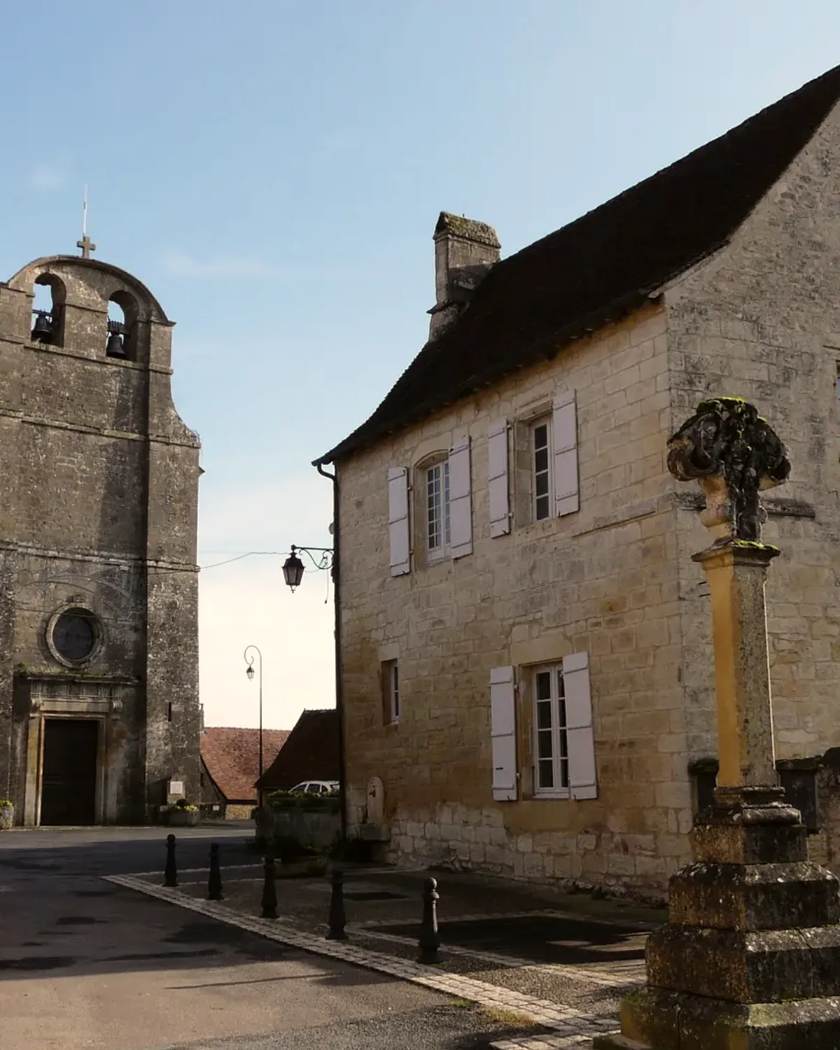 L'église de Fanlac, sa croix de pierre et les maisons de la place, sous un ciel bleu.