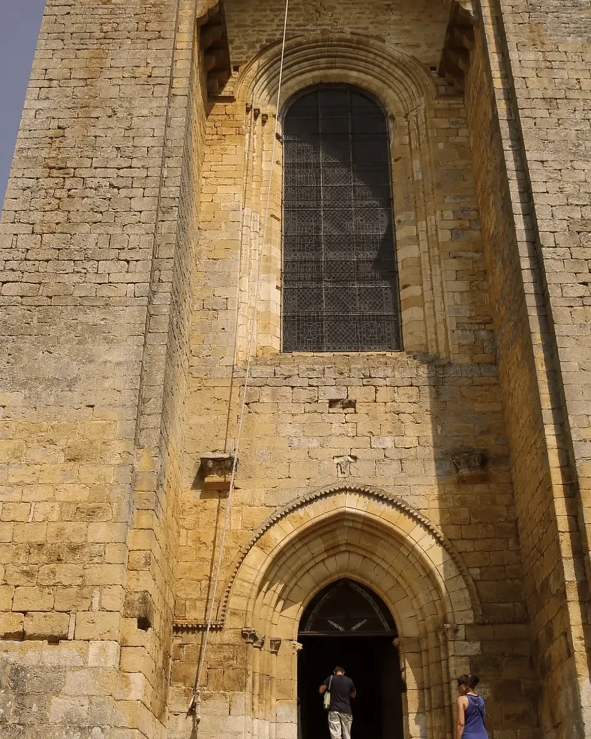 La façade de l'abbatiale fortifiée de Saint-Amand-de-Coly, deux visiteurs à l'entrée.